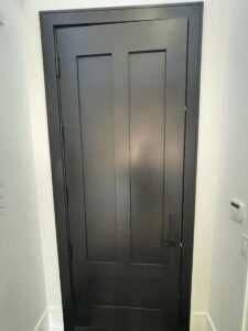 A closed, dark gray door with a simple, modern design. It features three vertical panels and is set in a white wall. A small light switch and a thermostat are visible on the right side of the frame.