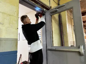 A person wearing gloves and a black and white jacket is using a power tool for door repair, focusing on the top of a metal door frame in an indoor setting. A concrete wall peeks through the scene, with tools scattered on the floor nearby.