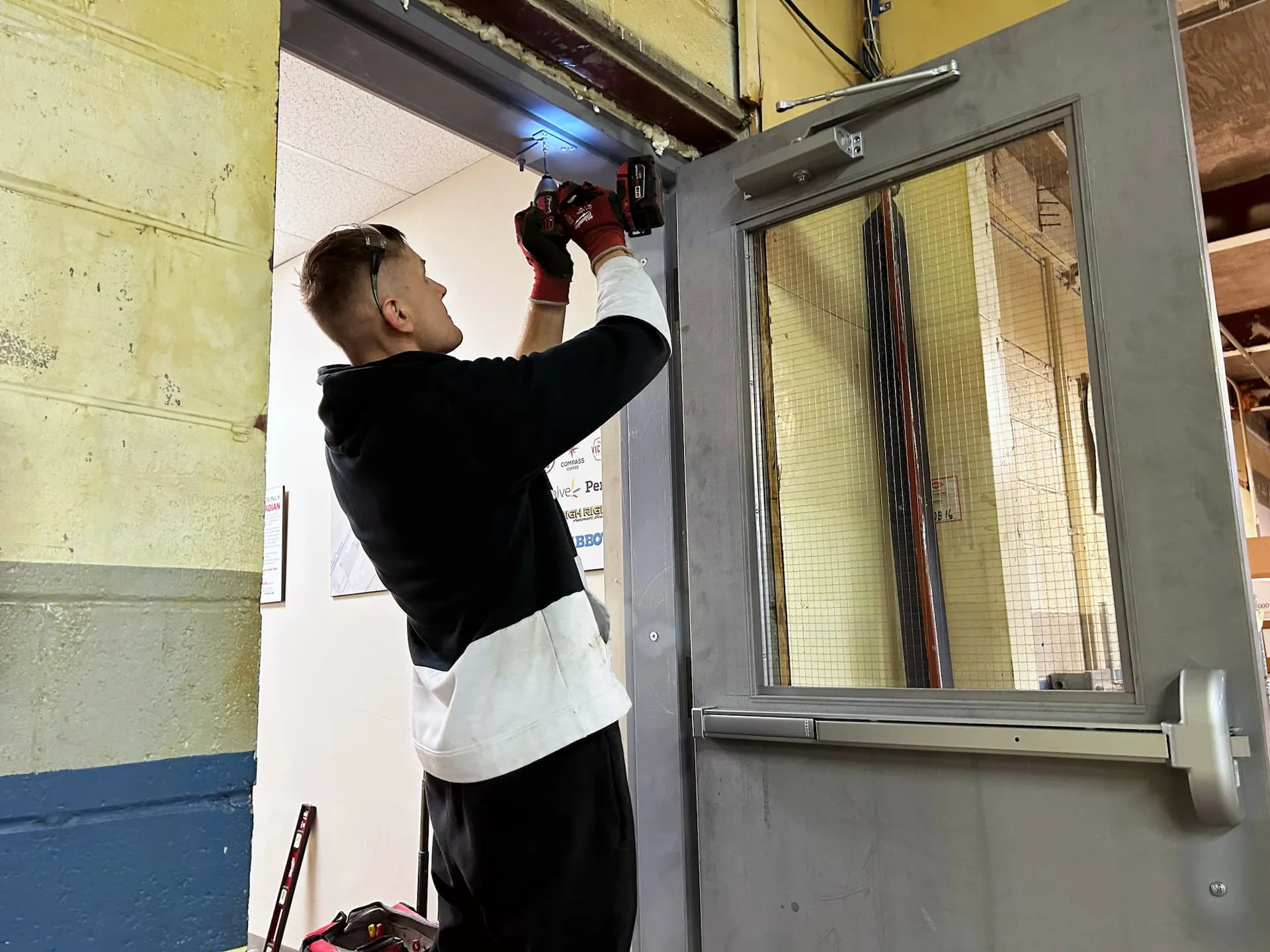 A person wearing gloves and a black and white jacket is using a power tool for door repair, focusing on the top of a metal door frame in an indoor setting. A concrete wall peeks through the scene, with tools scattered on the floor nearby.